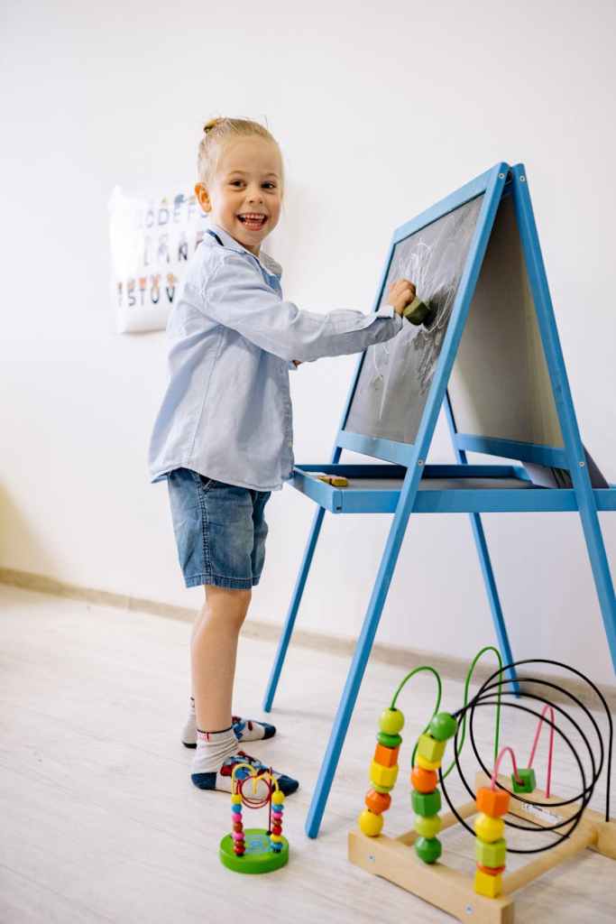 "Learning that belongs to the child stays with the child" attributed to John Holt, author of How Children Fail, over a background image of a child in a blue long sleeve shirt and blue denim shorts learning to draw on a blackboard.