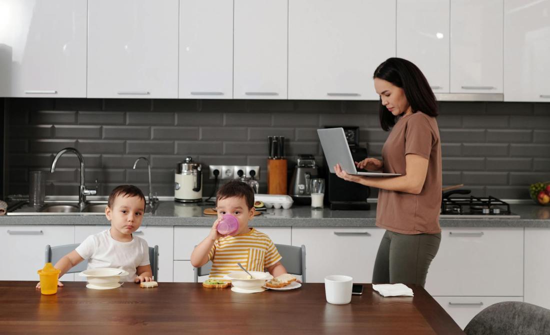 a woman using her laptop near her kids eating breakfast on a wooden table