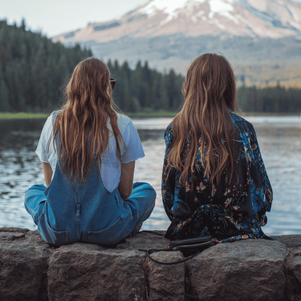 Two women sitting side by side on rocks beside a mountain lake — illustrating the difference between friendship and life coaching for homeschool moms.