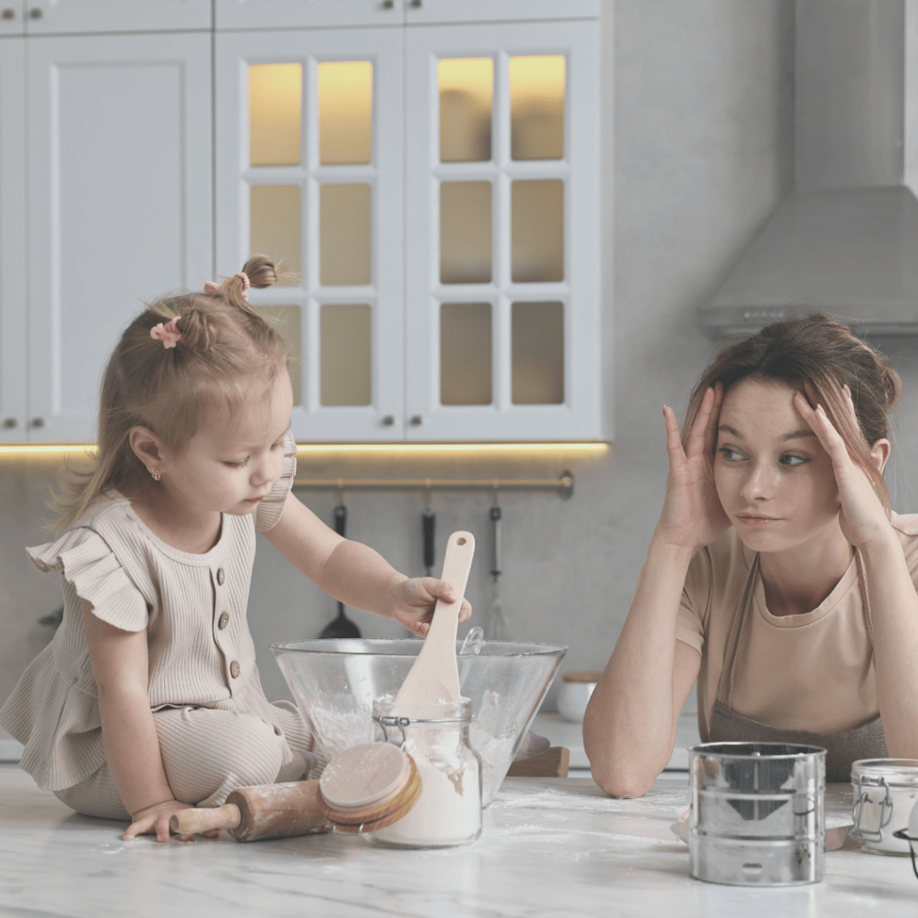 Overwhelmed homeschool mom sitting at the kitchen counter with her head in her hands while her young daughter bakes beside her